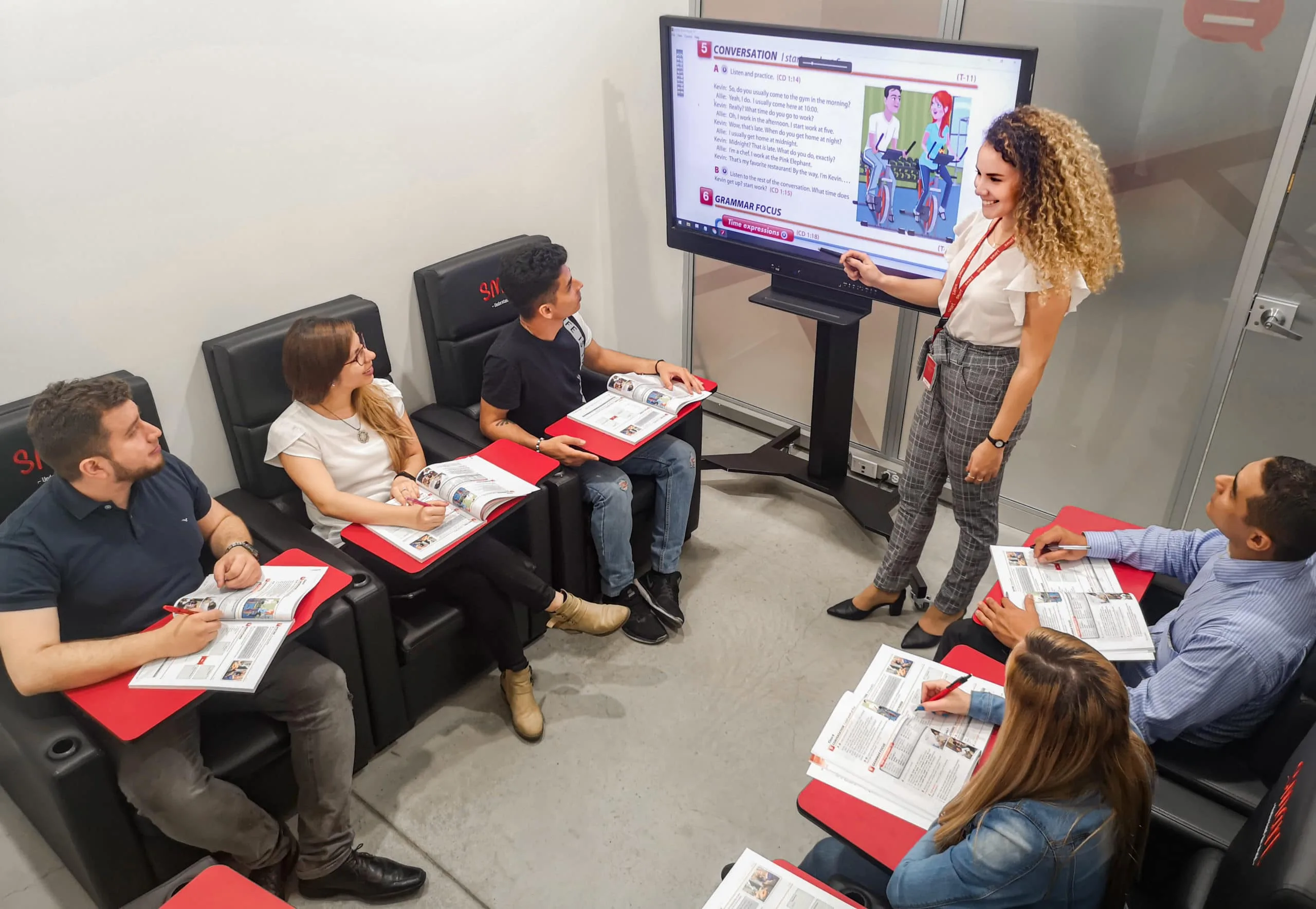 Docente de Smart junto a una pantalla con material de conversación frente a un grupo de estudiantes en clase